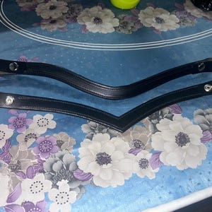 Pode incluir: Duas testeiras de couro preto com encaixes prateados. As testeiras têm formato em V e possuem um design costurado. O fundo é um padrão floral azul.