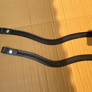 Puede incluir: Dos correas de cuero negro con cierres de botón a presión plateados. Las correas tienen un diseño curvo y están colocadas sobre una superficie de cartón marrón. Las correas parecen nuevas y sin usar.