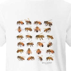 Puede incluir: Camiseta blanca con un patrón de ilustraciones detalladas de abejas. Las abejas son marrones y amarillas, con alas y detalles corporales visibles. El texto "MĀLA HONEY HAWAII" está impreso en la parte inferior.