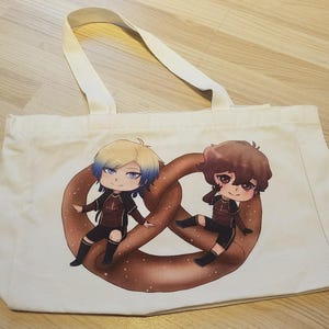 Könnte beinhalten: Eine weiße Stofftasche mit einem Grafikdruck von zwei Chibi-Figuren, die auf einer großen braunen Brezel sitzen. Die Figur auf der linken Seite hat blondes und blaues Haar, während die Figur auf der rechten Seite braunes Haar hat.
