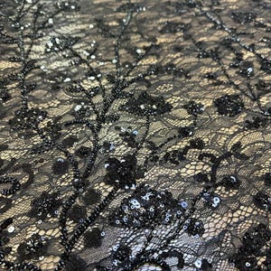 May include: Close-up of black lace fabric with intricate floral and beaded embellishments. The delicate lace features a floral pattern with beaded branches and scattered sequins, creating a textured and elegant design.