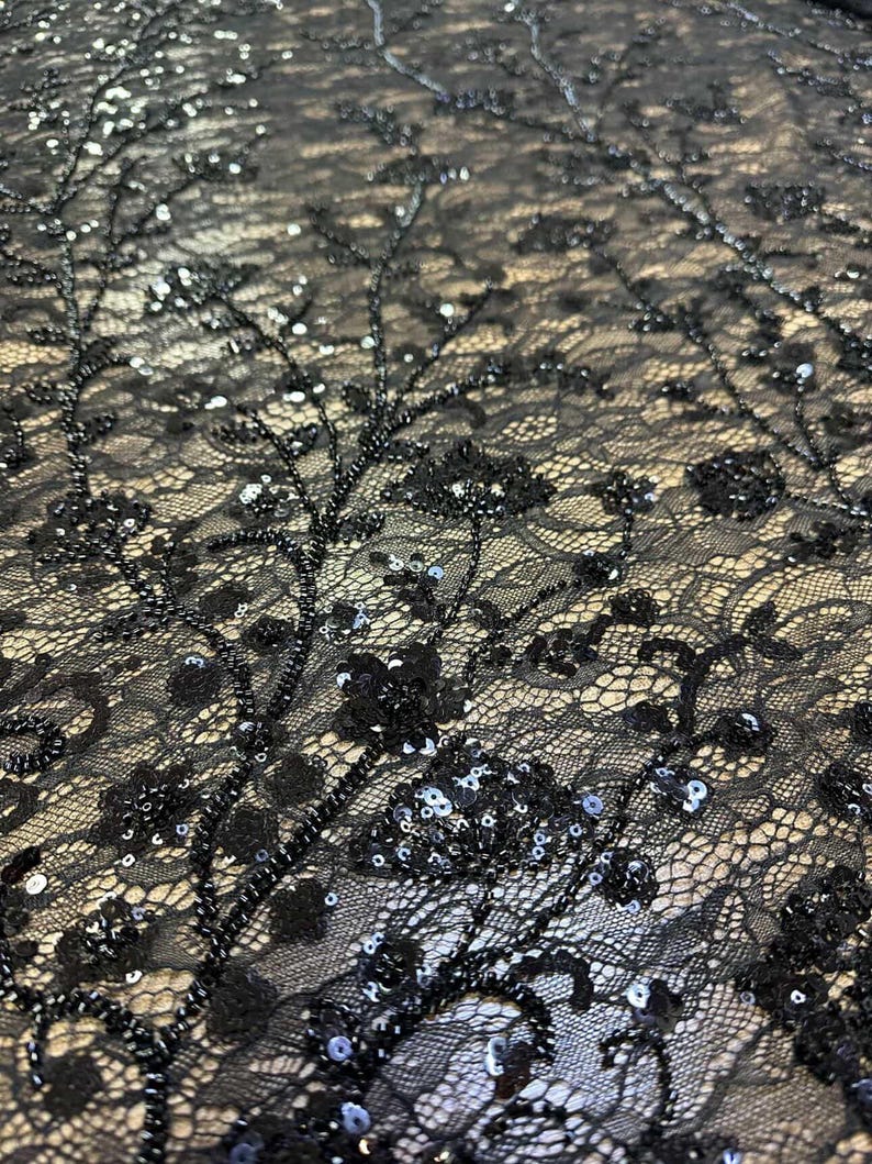 May include: Close-up of black lace fabric with intricate floral and beaded embellishments. The delicate lace features a floral pattern with beaded branches and scattered sequins, creating a textured and elegant design.