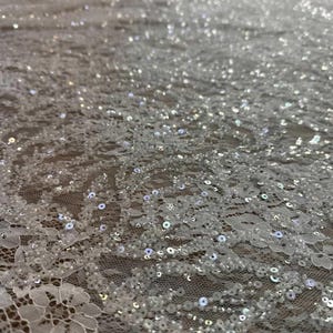 May include: Close-up of white lace fabric with intricate floral patterns and shimmering sequins. The delicate material is spread out, showcasing the detailed design and texture. Ideal for bridal wear or special occasion garments.