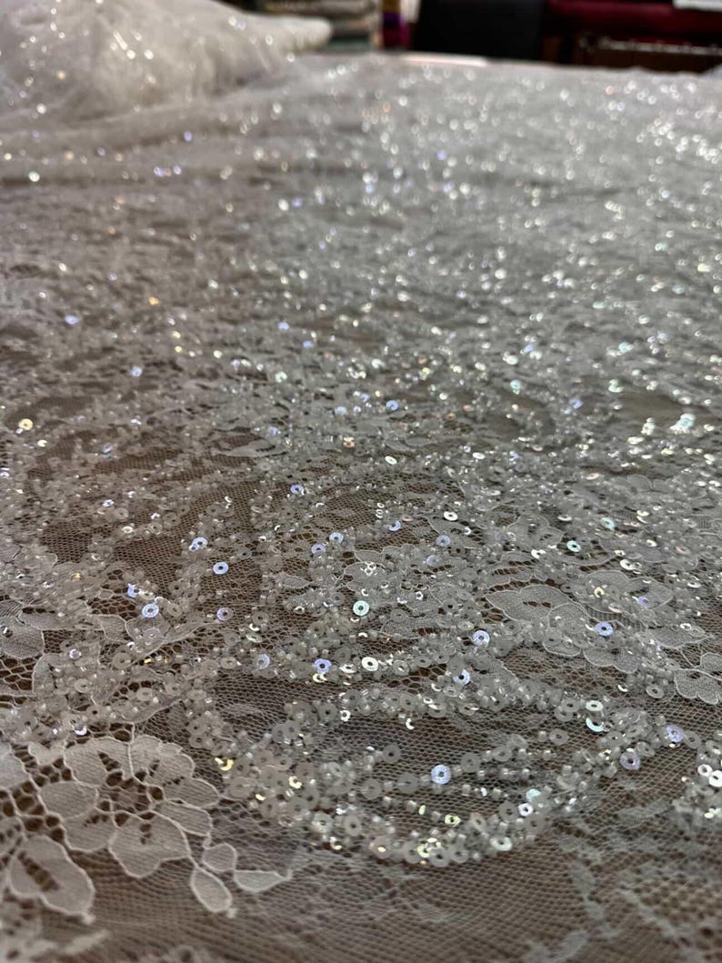 May include: Close-up of white lace fabric with intricate floral patterns and shimmering sequins. The delicate material is spread out, showcasing the detailed design and texture. Ideal for bridal wear or special occasion garments.