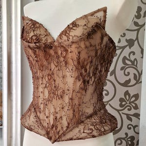 May include: A brown lace corset top with a sweetheart neckline and a structured, fitted silhouette. The delicate lace is embellished with small, scattered beads. The top is displayed on a white mannequin, showcasing its intricate design and craftsmanship.