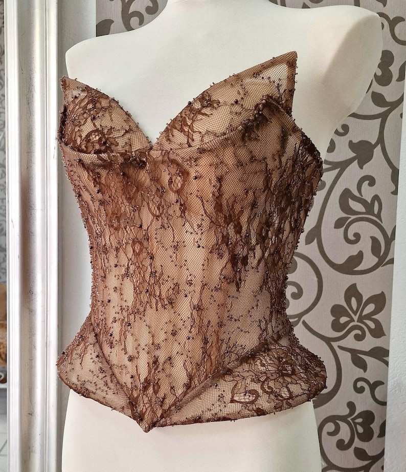 May include: A brown lace corset top with a sweetheart neckline and a structured, fitted silhouette. The delicate lace is embellished with small, scattered beads. The top is displayed on a white mannequin, showcasing its intricate design and craftsmanship.