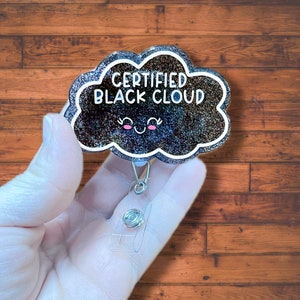 May include: A black glitter cloud-shaped badge holder with the text "CERTIFIED BLACK CLOUD" and a cute cartoon face. The badge holder is attached to a clear retractable badge reel.