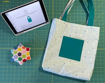 Puede incluir: Una bolsa de tela azul claro con un bolsillo verde y un estampado floral blanco. La bolsa tiene dos asas verdes. La bolsa está sobre una estera de corte verde con un alfiletero en forma de estrella blanca.
