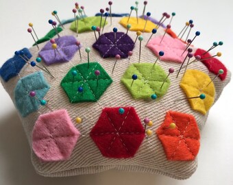 May include: A star-shaped pincushion with a beige fabric exterior and colorful felt hexagons. The hexagons are arranged in a pattern and are filled with pins.