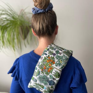 May include: A blue fabric scrunchie in a woman's hair. A white fabric neck wrap with a colorful floral print of potted plants is draped over the woman's shoulders.