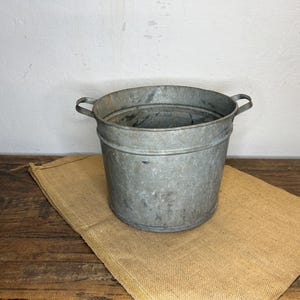 Op de afbeelding: Een gegalvaniseerde metalen emmer met twee handvatten, op een jute doek. De emmer is cilindrisch met een licht uitlopende rand. De achtergrond is een witte muur en een houten tafel.