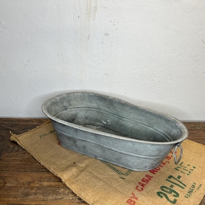 Può includere: Una vasca ovale in metallo zincato con due manici. La vasca è appoggiata su un sacco di juta con testo verde. La vasca è su una superficie di legno. Lo sfondo è un muro bianco.