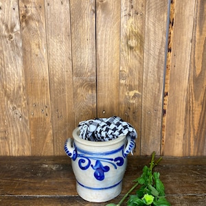 May include: A blue and white ceramic crock with two handles, filled with a black and white checkered fabric. The crock is sitting on a wooden surface with green flowers and a seed pod.