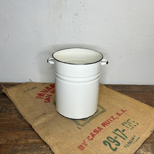 Op de afbeelding: Een witte emaille emmer met zwarte rand en twee kleine handvatten. De emmer staat op een jute zak met rode en groene tekst. De emmer is cilindrisch en heeft een eenvoudig, klassiek ontwerp, geschikt voor diverse doeleinden.