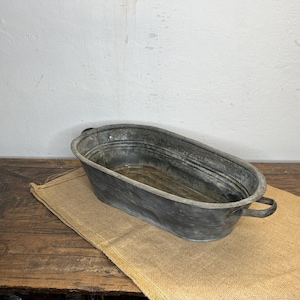 Può includere: Una vasca ovale in metallo zincato con due manici. La vasca è grigio scuro e poggia su un panno di juta beige. La vasca è su un tavolo di legno stagionato.