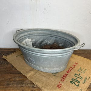 Könnte beinhalten: Ovale Zinkwanne mit zwei Griffen, auf einem Jutesack mit rotem und grünem Text. Die Wanne ist mit Erde gefüllt und steht auf einer Holzoberfläche. Ein Vintage-Behälter.