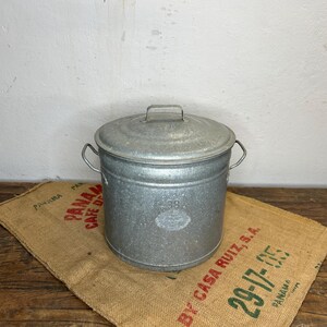 May include: A galvanized metal bucket with a lid and two handles. The bucket is sitting on a burlap sack with text. The bucket has a small handle on the lid and a number 38 on the front.