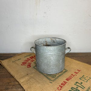 Può includere: Un secchio in metallo zincato con due piccole maniglie. Il secchio è appoggiato su un sacco di iuta con testo rosso e verde. Lo sfondo è una parete bianca e una superficie in legno.