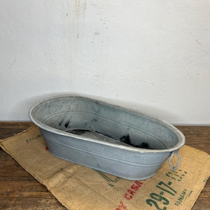 Può includere: Una vasca ovale in metallo zincato con manici, appoggiata su un sacco di juta. La vasca ha un aspetto usurato ed è appoggiata su una superficie di legno contro un muro bianco. Il sacco di juta ha testo verde.