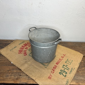 Op de afbeelding: Een ronde emmer van gegalvaniseerd metaal met twee handvatten aan de zijkant. De emmer heeft een gladde, zilvergrijze afwerking en een cilindrische vorm. Het staat op een beige koffiezak van jute met rode en groene bedrukte tekst.