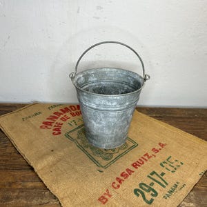 Könnte beinhalten: Ein verzinkter Metalleimer mit Drahtbügel steht auf einem Jutesack. Der Eimer hat eine abgenutzte, strukturierte Oberfläche. Der Jutesack hat roten und grünen Text und eine grüne Grafik.