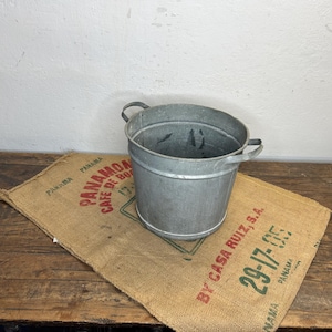 Op de afbeelding: Een cilindervormige emmer van gegalvaniseerd metaal met twee handvatten aan de zijkant, geplaatst op een beige koffiezak van jute. De zak bevat rood en groen gedrukte tekst, waaronder "PANAMA", "CAFE DE BOQUETE", "BY CASA RUIZ, S.A." en "29-17-05".