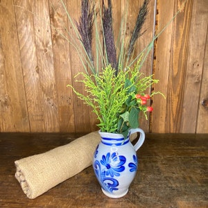 May include: A blue and white ceramic pitcher with a floral design, filled with dried grasses and red berries, sits on a wooden surface.