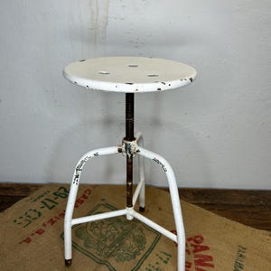 Op de afbeelding: Een vintage witte metalen verstelbare kruk met een ronde zitting en een driepoot. De kruk heeft een versleten, versleten uitstraling, met zichtbare afbladderende verf. De zitting heeft verschillende kleine gaten. De kruk staat op een jute zak.