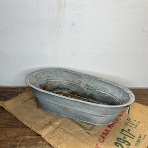 Può includere: Una vasca ovale in metallo zincato con bordi arrotolati e maniglie laterali. La vasca è appoggiata su un sacco di juta con testo sbiadito. Lo sfondo è un muro bianco e un tavolo di legno.