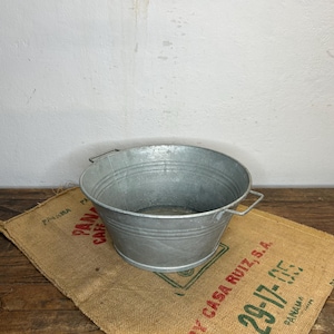 Può includere: Un secchio di metallo zincato con due manici è appoggiato su un sacco di juta con testo rosso e verde. Il secchio è rotondo con un bordo leggermente svasato e scanalature orizzontali. Il sacco di juta ha testo stampato e un bordo verde.