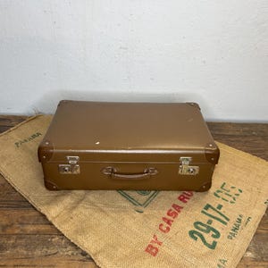 Può includere: Una valigia vintage marrone con chiusure e maniglia in metallo, appoggiata su un sacco di juta. La valigia ha angoli rinforzati e un design classico. Il sacco di juta ha testo stampato in rosso e verde.