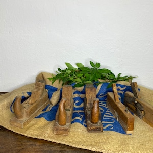 Könnte beinhalten: Fünf antike Holzhöbel auf einem Jutesack mit einem blauen und weißen Stoff darunter. Die Hobel sind in verschiedenen Größen und haben ein rustikales, abgenutztes Aussehen.