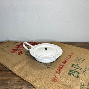 Op de afbeelding: Een witte po met deksel en handvat, geplaatst op een jute zak met rode en groene tekst. De po heeft een ronde vorm en een zwarte knop op het deksel. De zak ligt op een houten oppervlak.
