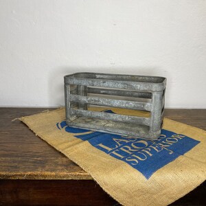 Könnte beinhalten: Ein Vintage-Flaschenkasten aus Metall mit rustikalem, verwittertem Finish. Der Kasten hat vier Fächer und steht auf einer Jutestoffe mit blauem Text, der "La Troya Superior" lautet.