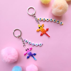 May include: Two metal keychains featuring beaded lizard charms and letter beads. One keychain has a yellow-to-red lizard with beads spelling DA BOMB. The other has a pink-to-blue lizard with beads spelling AS IF. Both include silver rings.