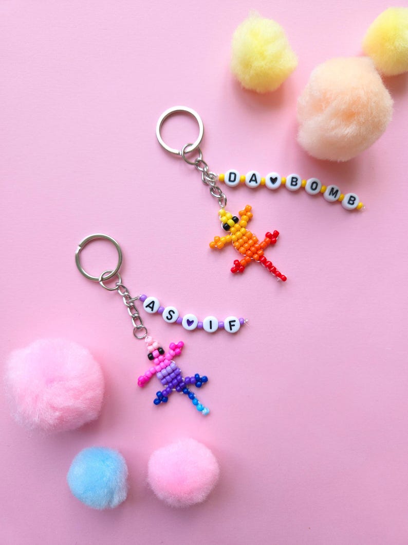 May include: Two metal keychains featuring beaded lizard charms and letter beads. One keychain has a yellow-to-red lizard with beads spelling DA BOMB. The other has a pink-to-blue lizard with beads spelling AS IF. Both include silver rings.