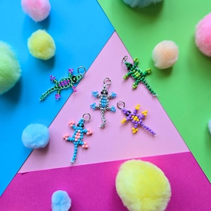 May include: Five lizard-shaped charms crafted from small colorful beads, each featuring a metal jump ring at the top. The lizards are designed in various color combinations including green and pink, light blue and pink, bright blue and white, green and yellow, and purple and yellow.