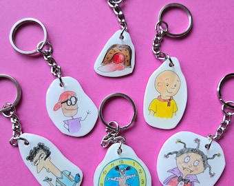 May include: Six white keychains featuring hand-drawn illustrations of various cartoon characters and objects, including a clock, a person in a yellow shirt, a person with glasses, a vampire-like figure, and a brown object with a red center.