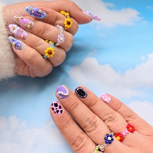 May include: A collection of beaded rings featuring colorful flower designs, including yellow, purple, blue, white, and red petals. The rings are worn on fingers with various nail art designs, including cow print, polka dots, and floral accents.