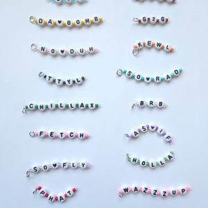 May include: A collection of small beaded charms featuring white alphabet beads with black letters and colorful accent beads. The charms display phrases like L8R, 143, DA BOMB, G2G, NO DUH, KEWL, TTYL, SO RAD, CHILLAX, BRB, FETCH, AS IF, SO FLY, HOLLA, PHAT, and WAZZZUP.