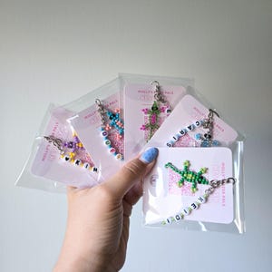May include: Five beaded lizard keychains in clear plastic packaging, each attached to a white card labeled Molly's Mozza Pals. The lizards are crafted from colorful seed beads, and each keychain features white letter beads spelling names.