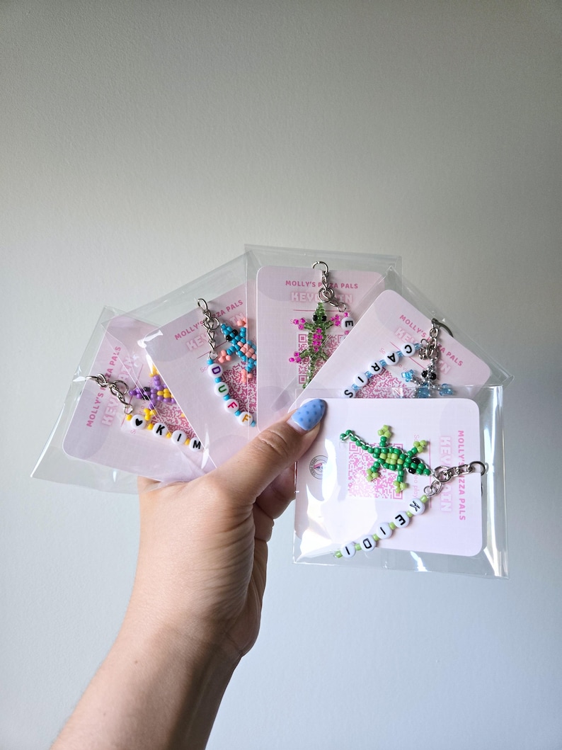 May include: Five beaded lizard keychains in clear plastic packaging, each attached to a white card labeled Molly's Mozza Pals. The lizards are crafted from colorful seed beads, and each keychain features white letter beads spelling names.