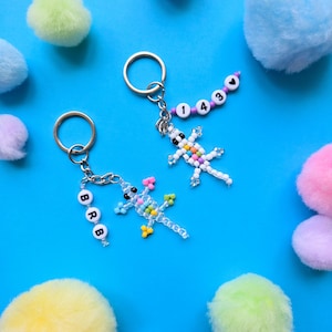May include: Two metal keychains featuring beaded lizard figures. One lizard is clear with colorful beads and a strand of white letter beads spelling BRB. The other lizard is white with colorful beads and a strand of purple beads with numbers 1430 and a heart.