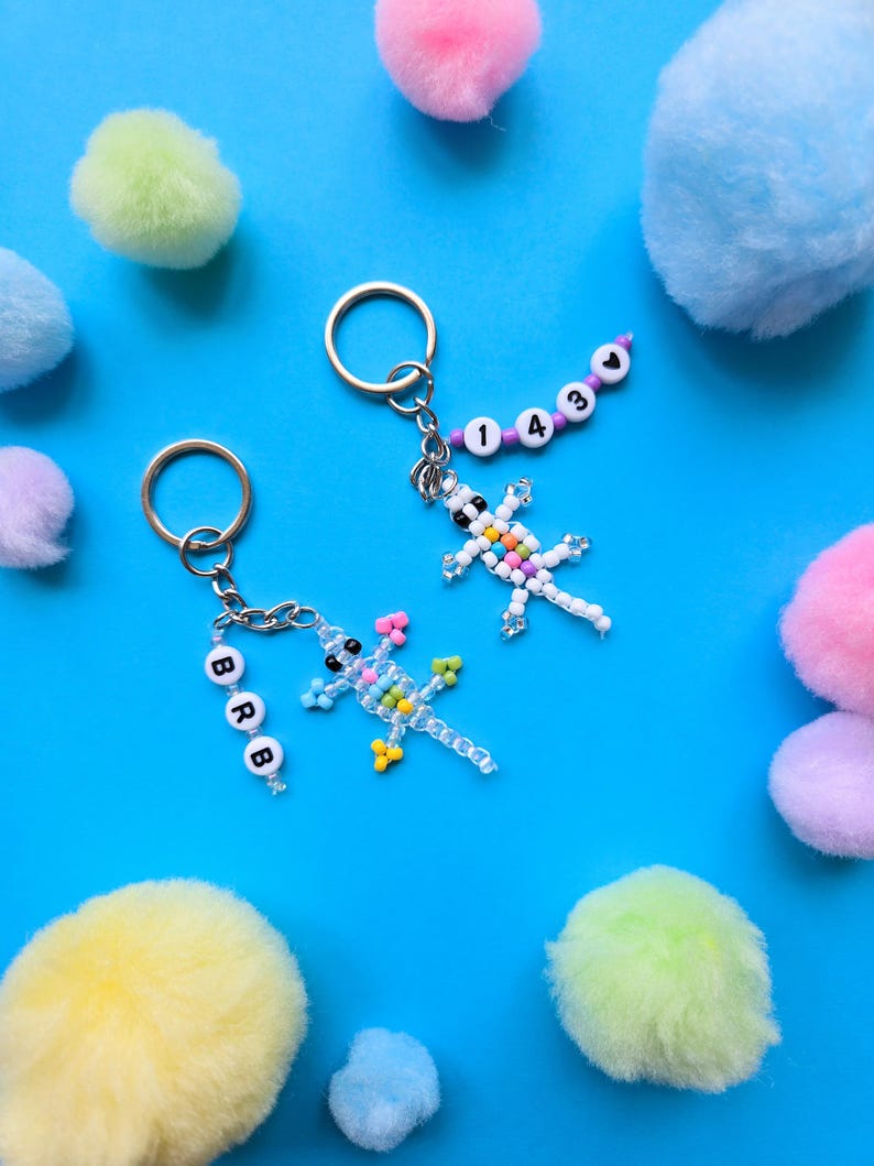 May include: Two metal keychains featuring beaded lizard figures. One lizard is clear with colorful beads and a strand of white letter beads spelling BRB. The other lizard is white with colorful beads and a strand of purple beads with numbers 1430 and a heart.