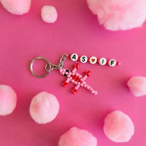 May include: A keychain featuring a beaded lizard in shades of pink and red, attached to a silver metal ring. A string of white beads with black letters spells out the name A S O I F, with a small red heart bead replacing the letter O.