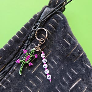 May include: A black textured zipper pouch featuring a metal keychain attached to the zipper pull. The keychain includes a green and pink beaded lizard charm and a vertical strand of white letter beads spelling out the name EMILY.