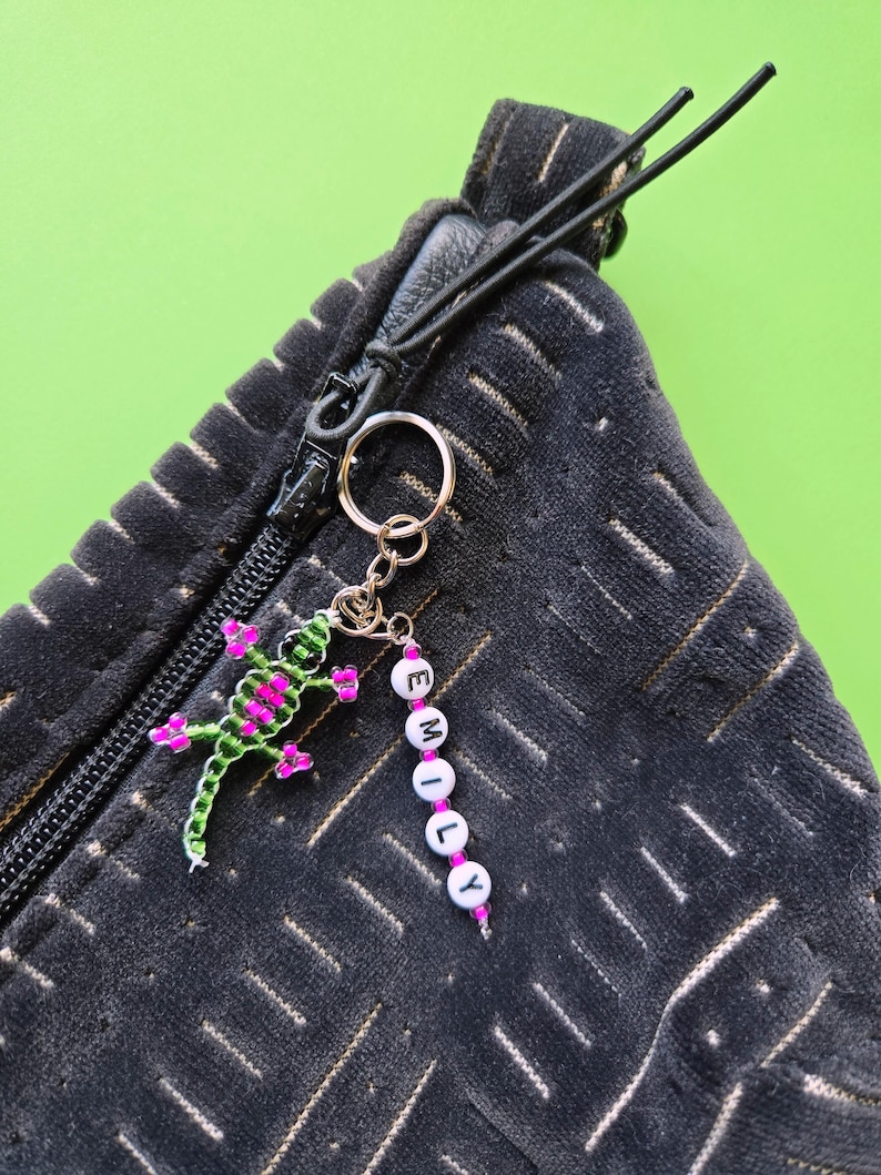 May include: A black textured zipper pouch featuring a metal keychain attached to the zipper pull. The keychain includes a green and pink beaded lizard charm and a vertical strand of white letter beads spelling out the name EMILY.