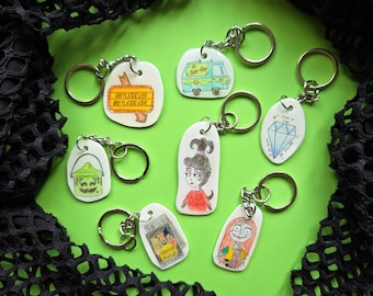 May include: A collection of seven white acrylic keychains featuring various pop culture illustrations, including a Betelgeuse sign, a mystery van, a diamond with text, a green bucket, a cartoon character, a Hocus Pocus bag, and a Sally character.