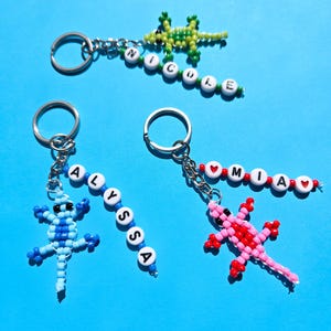 May include: Three beaded lizard keychains with metal rings. One green lizard with white beads spelling NICOLE, one blue lizard with white beads spelling ALYSSA, and one pink and red lizard with white beads spelling MIA with red heart beads.