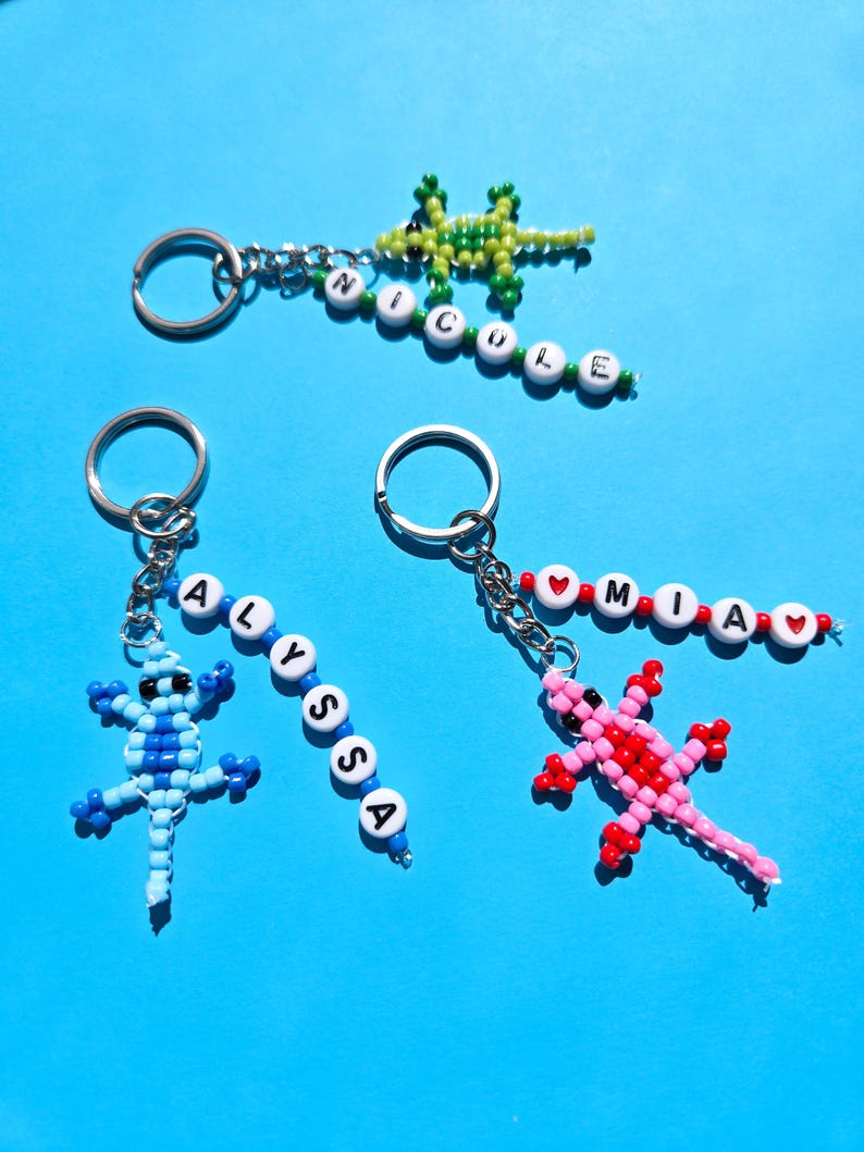 May include: Three beaded lizard keychains with metal rings. One green lizard with white beads spelling NICOLE, one blue lizard with white beads spelling ALYSSA, and one pink and red lizard with white beads spelling MIA with red heart beads.